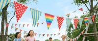 Children playing outdoors at summer party with colorful triangle bunting flags hanging between trees in backyard celebration setting.