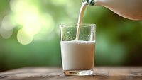 Fresh milk being poured into glass on wooden surface with natural green bokeh background for healthy breakfast and dairy nutrition concepts.