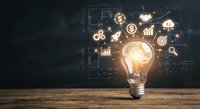 Glowing light bulb with financial icons and business symbols floating around it on wooden desk against dark background with mathematical formulas.