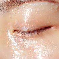 Close-up of closed eye with glossy, dewy skin effect and sparkle highlights, showing natural eyelashes and eyebrow detail against peachy complexion. Perfect for beauty and skincare.