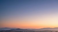 Serene mountain landscape at dawn with soft gradient sky transitioning from deep blue to warm orange and pink hues over silhouetted hills and valleys.