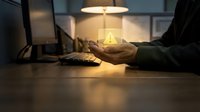 Hands holding glowing light bulb at desk with laptop, representing innovation, ideas, and sustainable energy solutions for business and technology concepts.