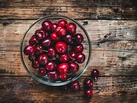 Fresh ripe cherries in glass bowl on rustic wooden background. Top view of sweet summer berries with stems for healthy eating and food design.