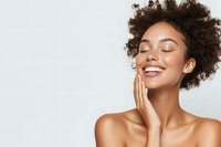 Young African American woman with natural curly hair and glowing skin smiling with eyes closed, touching face gently against light background. Wellness and skincare concept.