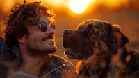 Young Caucasian man with curly hair and sunglasses sharing joyful moment with Australian Shepherd mix dog during golden hour sunset, creating warm emotional connection.