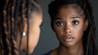 Young African American woman with natural dreadlocks and expressive eyes gazing at reflection in mirror against dark background, dramatic studio lighting highlights facial features.