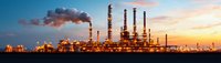 Panoramic view of oil refinery plant with illuminated industrial infrastructure and smoking chimneys against dramatic sunset sky with orange clouds.