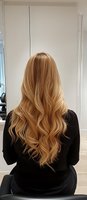 Woman with long blonde wavy hair sitting in modern hair salon chair wearing black top, rear view showcasing professional hairstyling results.