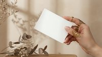 Female hand with ornate ring holding blank white card mockup against neutral beige background with dried botanical shadows for branding design.