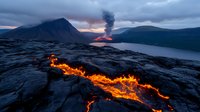 Dramatic volcanic eruption with flowing lava stream across black rock landscape, mountain silhouette and smoke plume against moody twilight sky over calm water.
