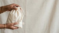 Woman with medium skin tone holding open a natural cotton drawstring bag against neutral linen background. Suitable for eco-friendly, sustainable lifestyle, and zero-waste shopping concepts.