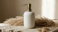 White ceramic soap dispenser with brass pump on natural linen fabric with dried pampas grass. Modern bathroom accessory for luxury spa and home decor styling.