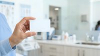 Close-up of male hand holding blank white business card in modern office bathroom setting with mirror and sink visible in background.