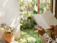 Close-up of hands holding open blank notebook by sunny window with potted plant and garden view, suitable for education, reading, writing, journaling, and lifestyle content.