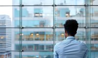 Young African American businessman looking through office window at urban cityscape, rear view shot showing contemplative moment in corporate environment.
