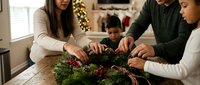 Mixed-race family creating festive holiday wreath together at home table with evergreen branches, berries and decorations during Christmas season.