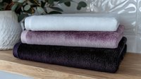 Stack of folded soft cotton bath towels in white, dusty pink and deep purple colors arranged on wooden surface against light background. Suitable for spa and bathroom decor.
