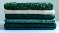 Stack of folded green and white textiles in various textures including knitted wool sweaters and cotton fabrics for cozy home decor and fashion concepts.