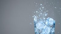 Glass of cold water with ice cubes splashing and dispersing against neutral gray background, suitable for beverage, refreshment, and hydration concepts.