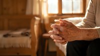 Elderly hands clasped in prayer or meditation in warm sunlit room with wooden furniture and peaceful atmosphere for spiritual wellness concepts.