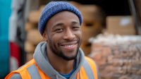 Young African American male worker wearing safety vest and blue beanie smiles warmly while working outdoors at industrial or construction site, expressing positivity and confidence.