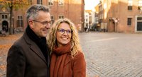 Happy mature couple embracing in historic European city square during golden hour sunset, wearing glasses and autumn coats, romantic travel lifestyle.