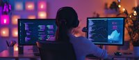 Young Asian female professional working late at computer monitors displaying data analytics and code in modern office with ambient purple lighting and decorative elements.