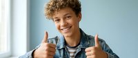 Happy mixed-race teenage boy with curly hair giving thumbs up gesture while smiling at camera in casual denim jacket for youth lifestyle content.