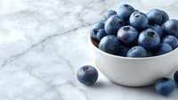 Fresh blueberries in white bowl on marble surface with scattered berries for healthy eating and cooking recipes.