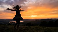 Woman with long dark hair in silhouette, dancing freely on hilltop at golden sunset with arms outstretched, celebrating nature and freedom outdoors.