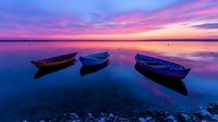 Three wooden boats floating on calm water reflecting vibrant purple and pink sunset sky, creating mirror-like surface with dramatic twilight colors.