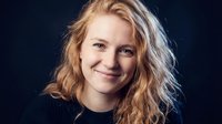 Young blonde Caucasian woman with wavy hair smiling warmly at camera against dark background. Professional headshot portrait for business and lifestyle use.