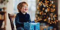 Happy young boy with blonde hair laughing while holding blue polka dot gift box during Christmas morning celebration with decorated tree lights.