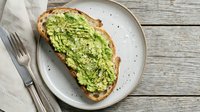 Avocado toast with seeds on rustic sourdough bread served on white ceramic plate with fork on wooden table background for healthy breakfast menu.