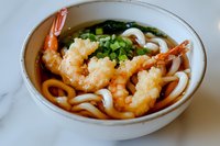 Tempura shrimp udon noodle soup in ceramic bowl garnished with fresh green onions and served in savory dashi broth on white marble surface.