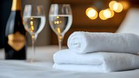 Clean white spa towels with champagne glasses and bottle in luxury hotel room setting, soft bokeh lights create romantic ambiance for wellness retreat.