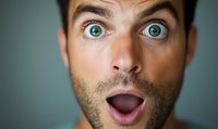 Closeup portrait of surprised young Caucasian man with bright blue eyes and beard showing shocked facial expression with open mouth against blurred background.