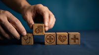 Male hand placing wooden block with dollar sign among healthcare and medical symbols on blue background, representing healthcare costs and medical expenses.