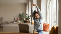 Young African American woman stretching arms up while working from home office, taking break from laptop computer in bright modern living room.