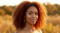 Young African American woman with curly hair smiling outdoors during golden hour sunset with warm natural lighting and blurred autumn background.