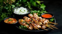 Grilled chicken skewers with sesame seeds served on black plate alongside spicy Asian dipping sauces and fresh herbs against dark background. Food photography.