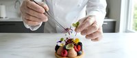 Pastry chef decorating gourmet dessert with fresh berries and edible flowers using tweezers in professional kitchen setting.