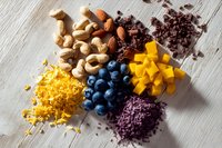 Assorted healthy ingredients on wooden background including nuts, blueberries, mango cubes, chocolate nibs, grated cheese, and purple rice for nutritious meal preparation.