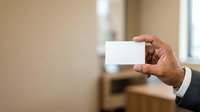 Close-up of male hand in dark business suit holding blank white business card against blurred office background, suitable for corporate branding and professional networking materials.