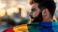 Young bearded man with pride rainbow flag wrapped around shoulders gazes thoughtfully at sunset cityscape, celebrating diversity and inclusion at pride event.