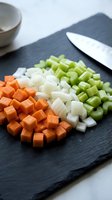 Fresh diced vegetables on slate cutting board with knife for cooking preparation including carrots onions and celery ready for soup or stew recipes.