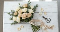 Elegant bridal bouquet with cream roses and eucalyptus on white wooden background with scattered petals and vintage scissors for wedding planning.