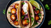 Crispy fish tacos garnished with red cabbage slaw, fresh cilantro and lime wedges on corn tortillas served on dark plate with vibrant toppings and creamy sauce.