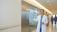Female doctor in white coat presenting medical heart scan on digital display screen in modern hospital corridor for healthcare technology presentation.