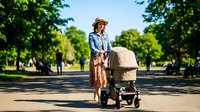 Woman in casual vintage attire with straw hat pushing baby carriage down tree-lined park path on sunny day. Suitable for parenting, lifestyle, and outdoor leisure concepts.